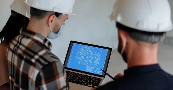 Trois professionnels du chantier consultent un plan sur un ordinateur portable.